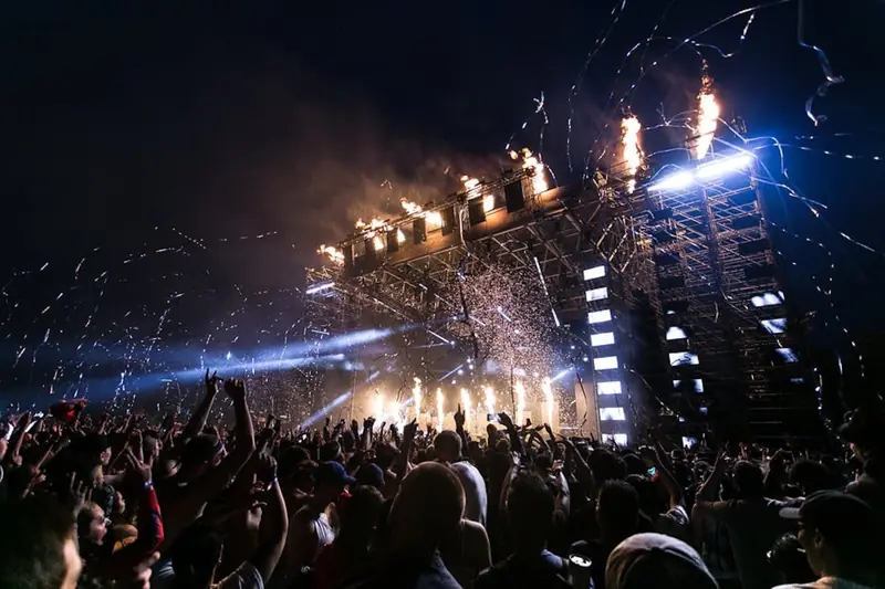 A festival crowd with hands up as pyrotechnics fire over a massive night stage